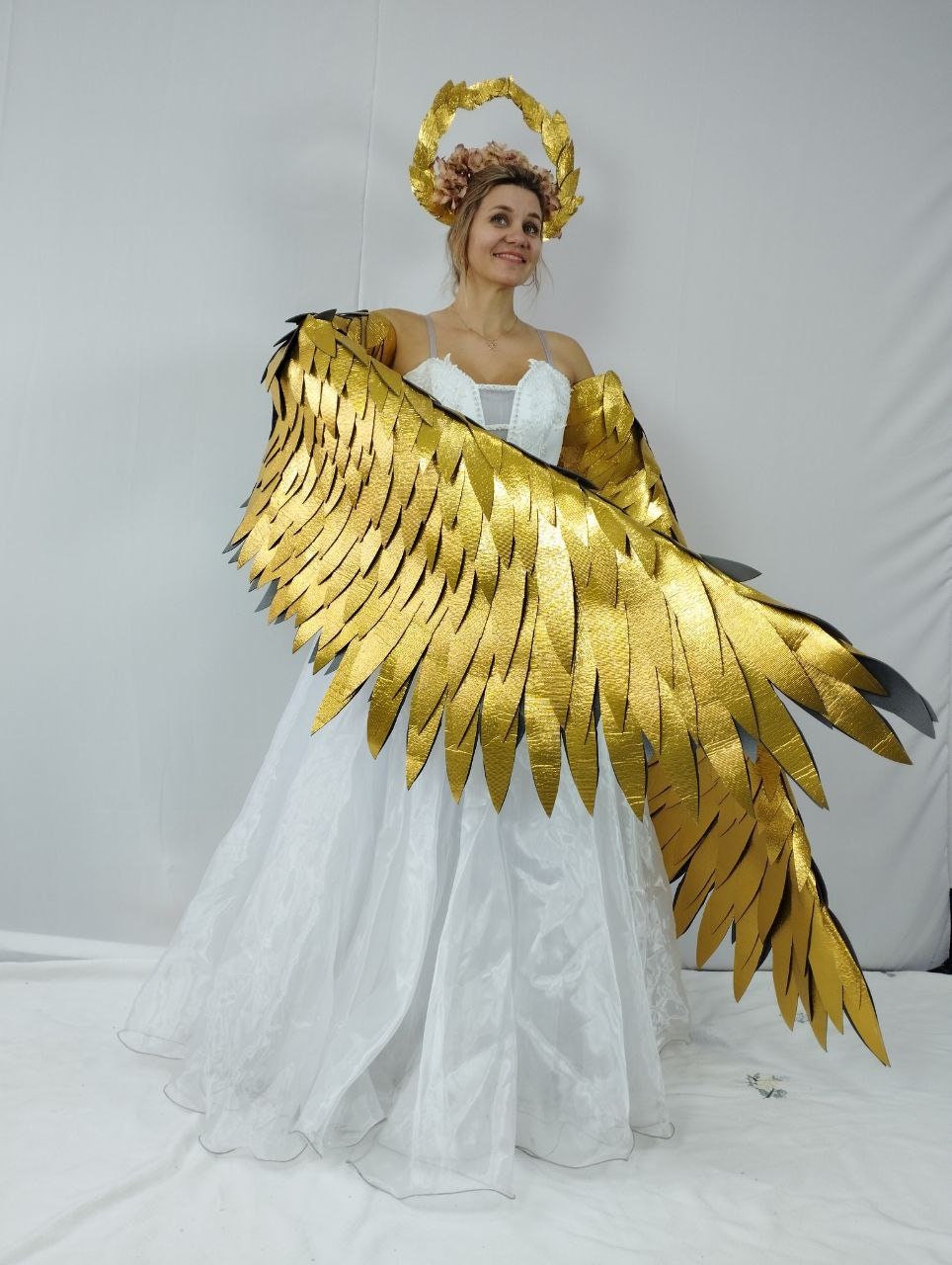 May include: A person wearing a white dress and gold-colored angel wings. The wings are large and detailed, with a layered design. A gold-colored halo with pink flowers sits atop the head. The dress is white and flowing, creating a celestial theme.