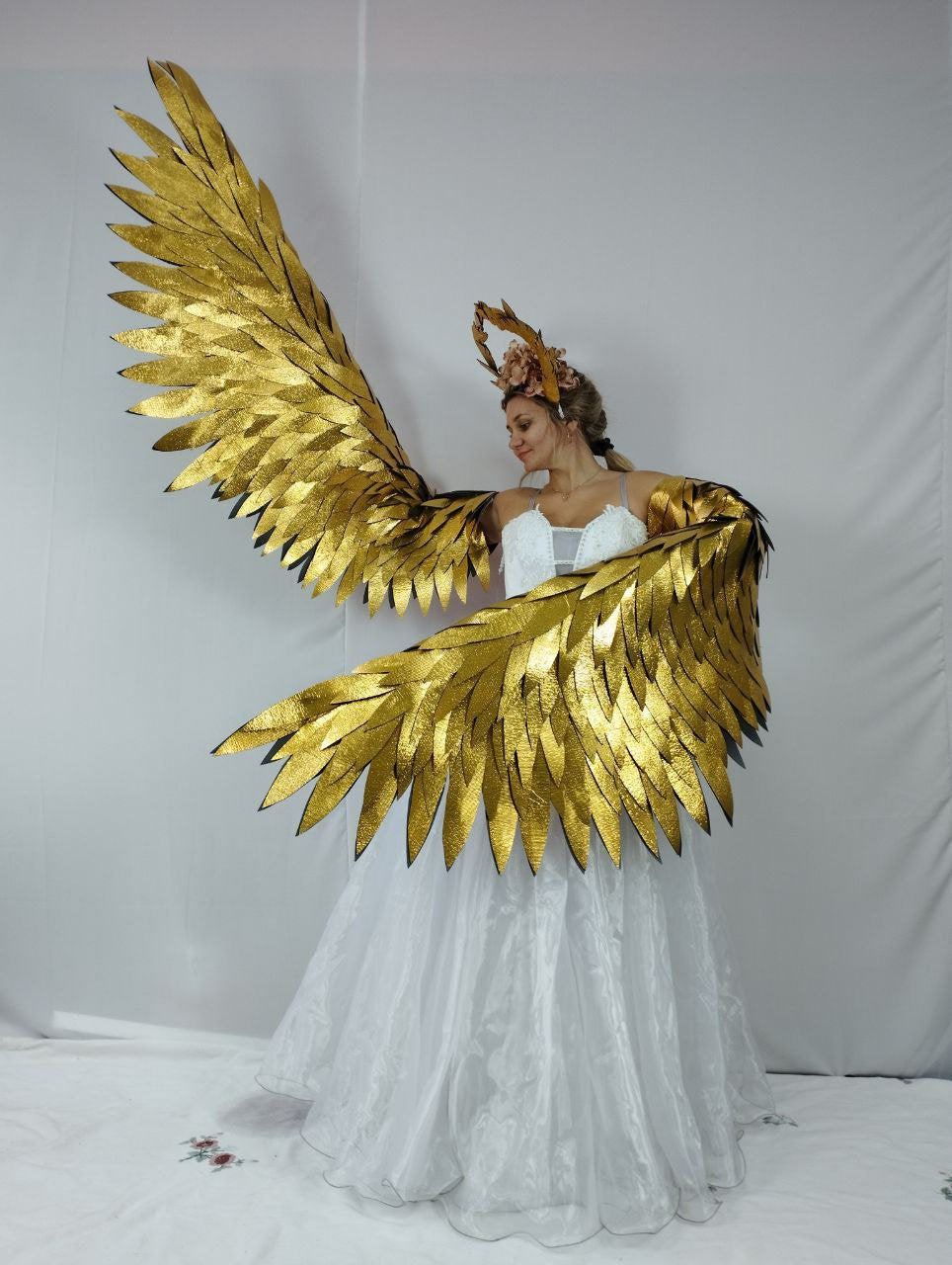 May include: A woman wearing a white dress and a gold-colored wing costume. The wings are large and detailed, with a layered feather design. A matching gold headpiece with floral accents completes the look. The costume is likely for a theatrical performance or cosplay.