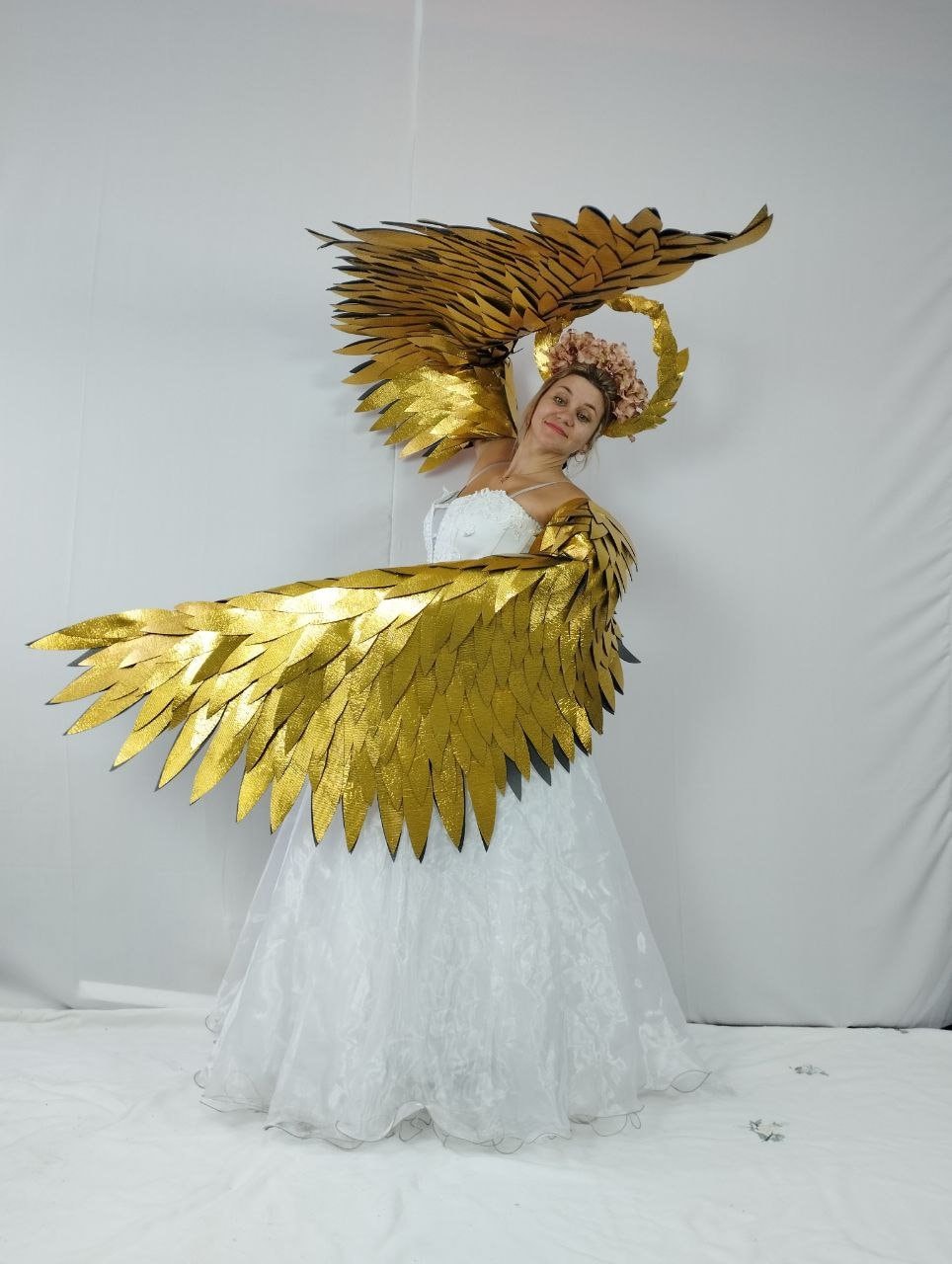 May include: A woman wearing a white dress and large gold-colored angel wings. The wings are detailed with layered feathers. A floral crown and halo complete the angelic costume. The wingspan is approximately 60 inches.