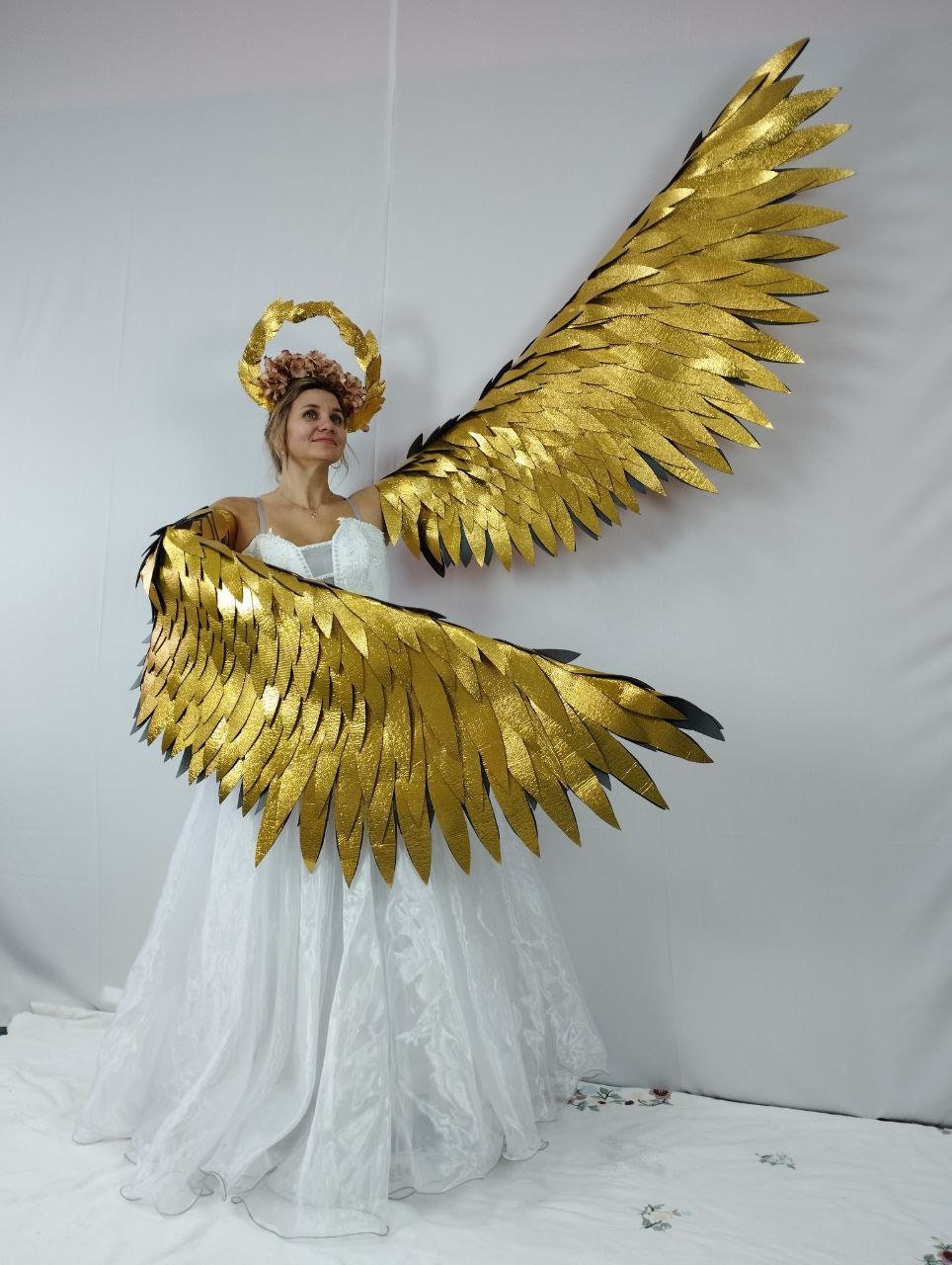 May include: A woman wearing a white dress, a gold halo, and large, gold, feathered wings. The wings are spread wide, and the woman is smiling. The wings are made of many individual pieces, giving them a textured appearance.