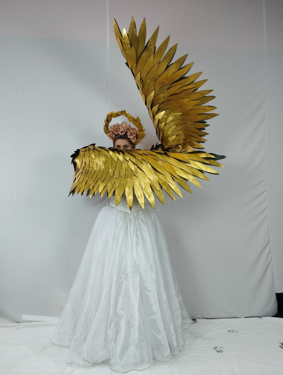 May include: A person wearing a gold-colored angel wing costume with a white dress and a floral crown. The wings are large and detailed, with a layered design. The costume is set against a white backdrop.