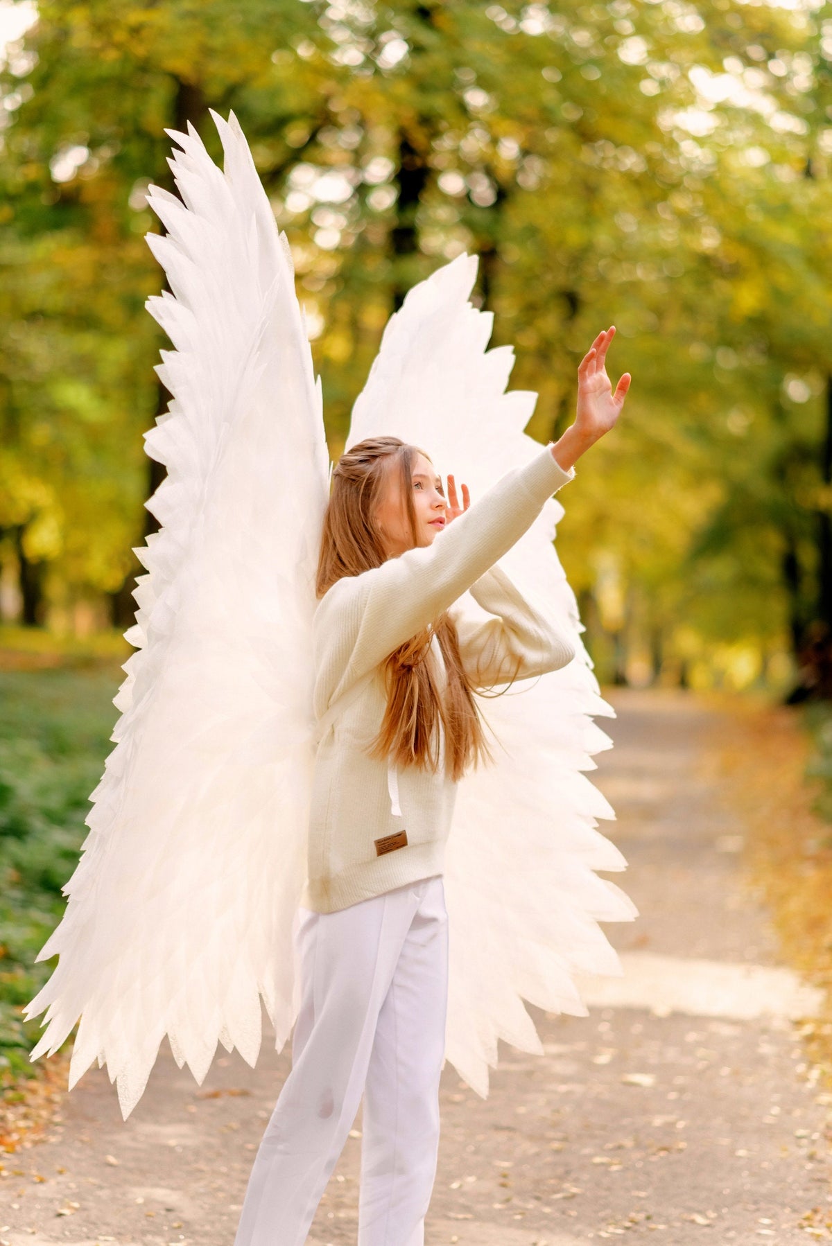 May include: A young person wearing a white sweater and white pants with large white angel wings. The person is standing in a forest with fall foliage in the background.