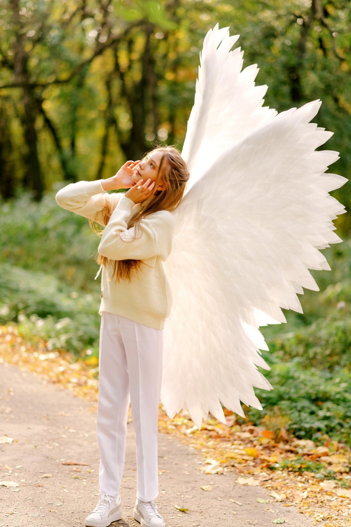 May include: A young person wearing a white sweater and white pants with large white angel wings standing in a park with fall leaves on the ground.