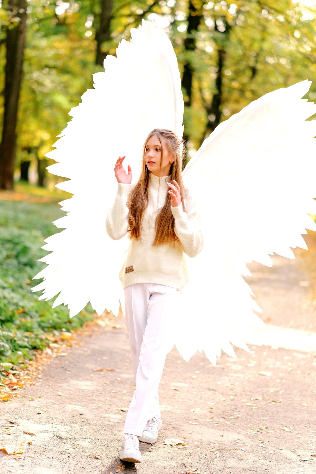 May include: A young person wearing a white sweater and white pants with large white angel wings in a park setting.