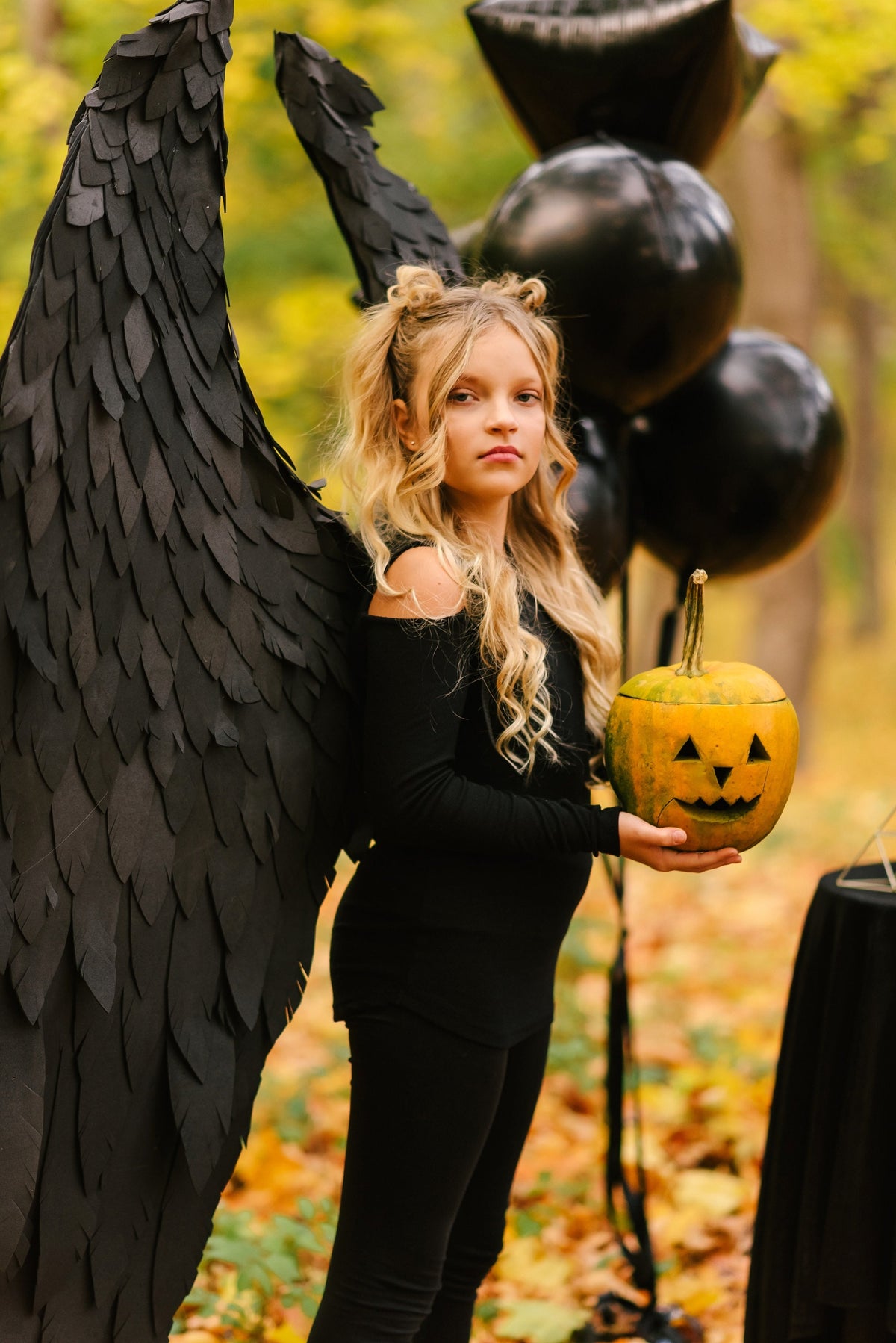 May include: A young girl dressed in black with black angel wings holds a carved pumpkin in a fall setting.