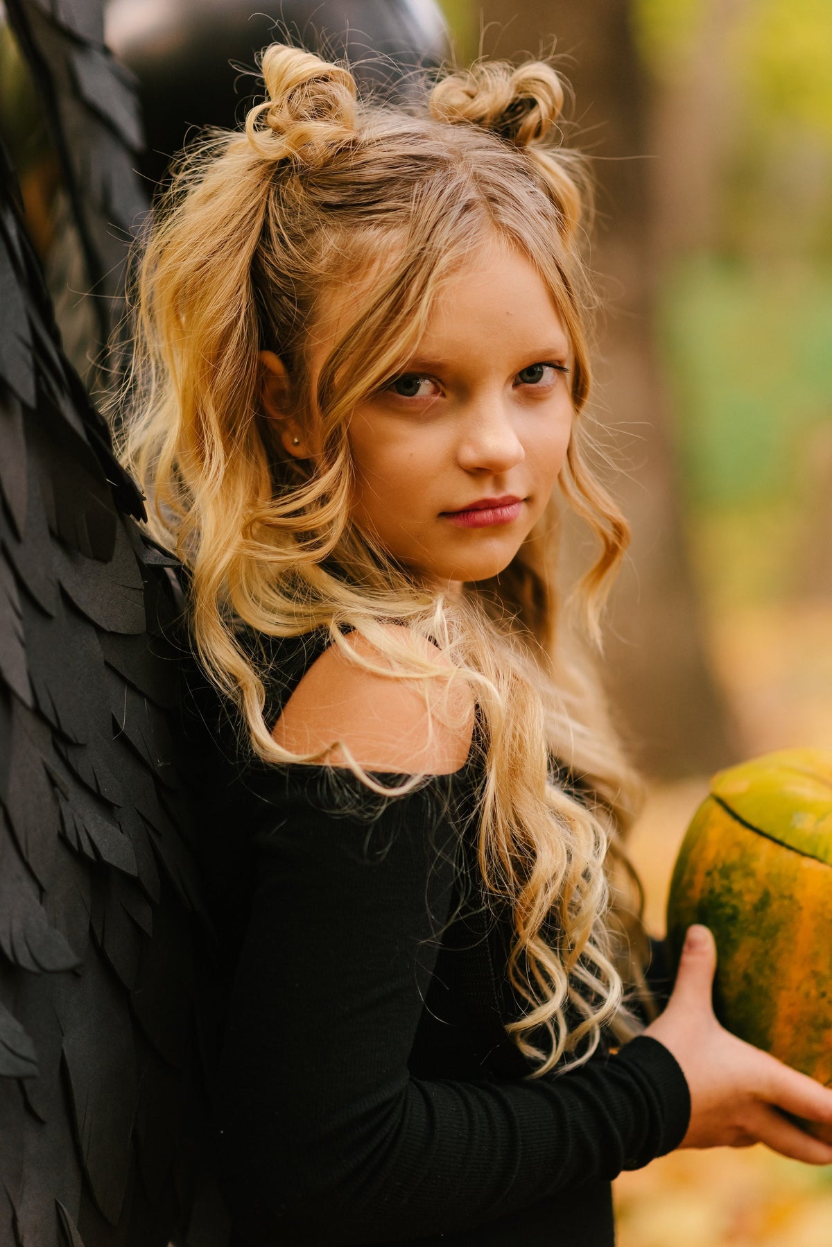 May include: A young girl with blonde hair styled in two pigtails with black bows, wearing a black off-the-shoulder top, holds a green and orange pumpkin.