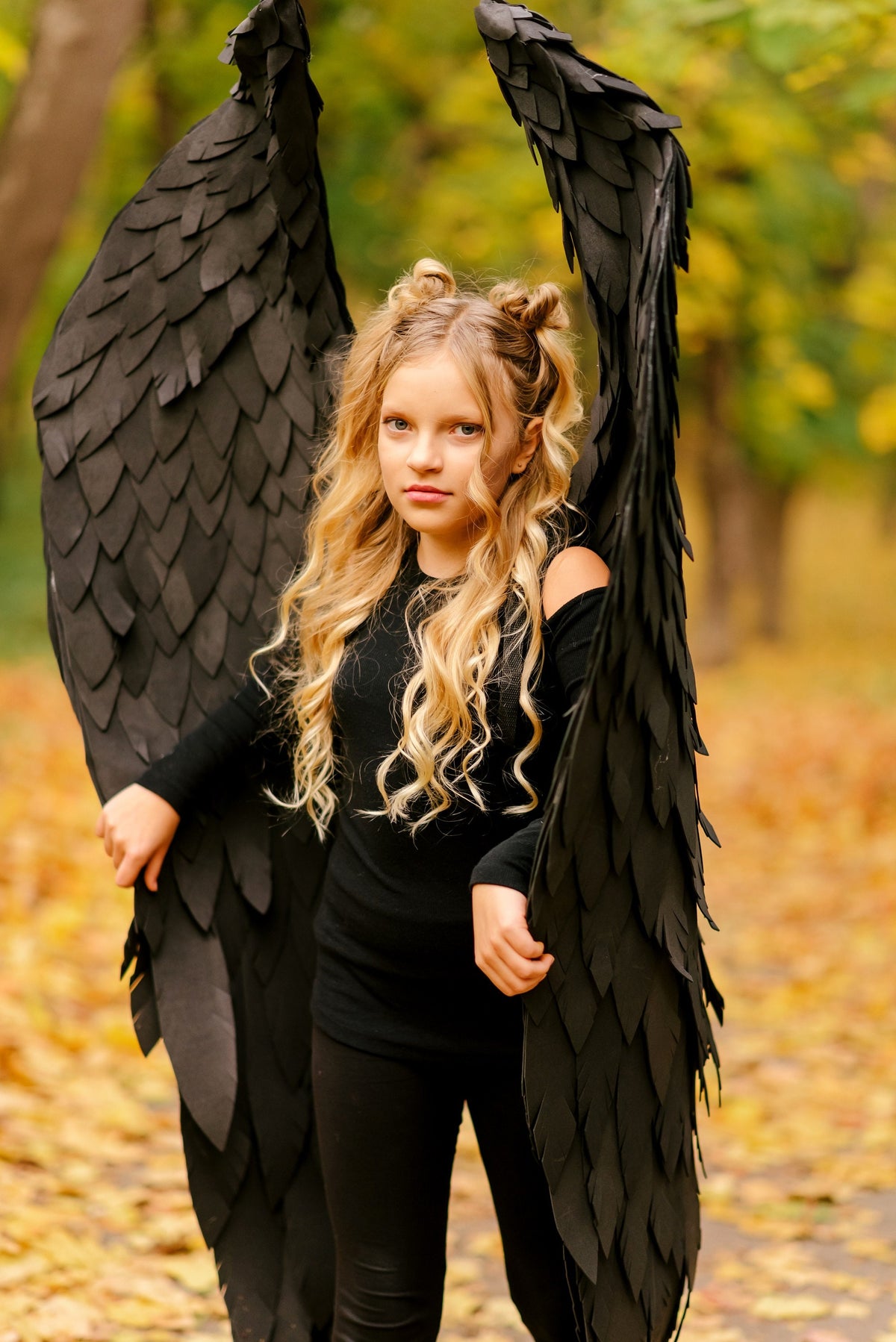 May include: A young girl with long blonde hair wearing a black outfit and large black wings. She is standing in a forest with fallen leaves on the ground.