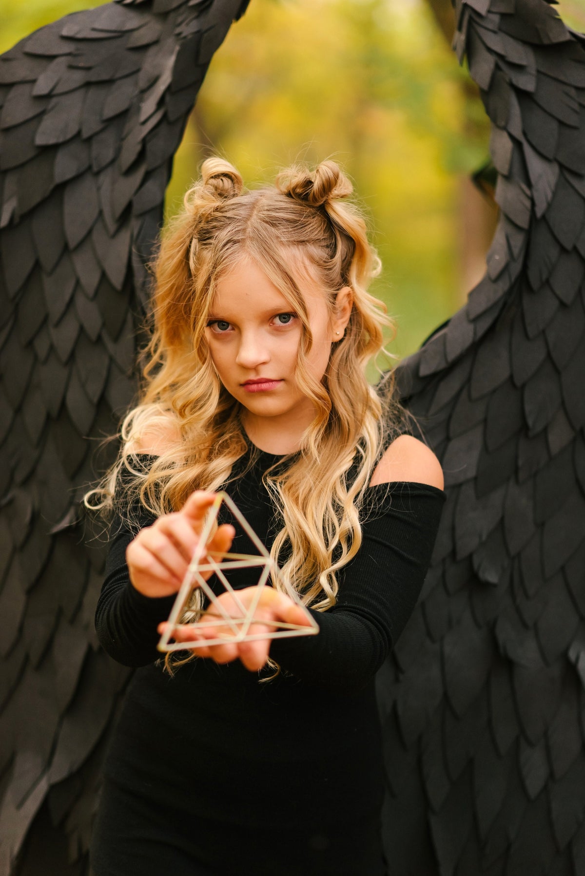 May include: A young person with long blonde hair wearing a black dress and black angel wings holds a gold geometric shape in front of them.