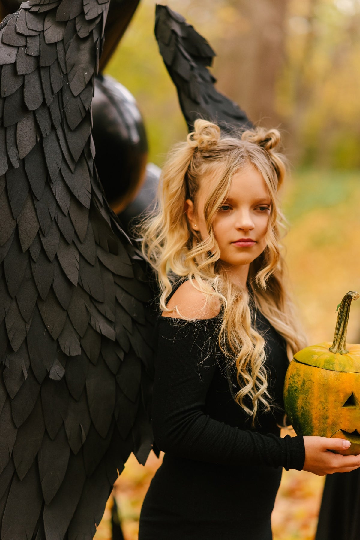 May include: A young girl dressed in black with black angel wings holds a carved pumpkin. She has blonde hair styled in two pigtails. The background is a blurry image of fall foliage.