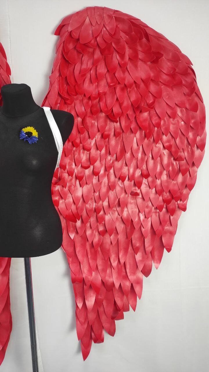 May include: A pair of large, vibrant red angel wings with a layered feather design. The wings are displayed next to a black mannequin torso with a white strap and a blue and yellow flower pin. The wings are made of a flexible material.
