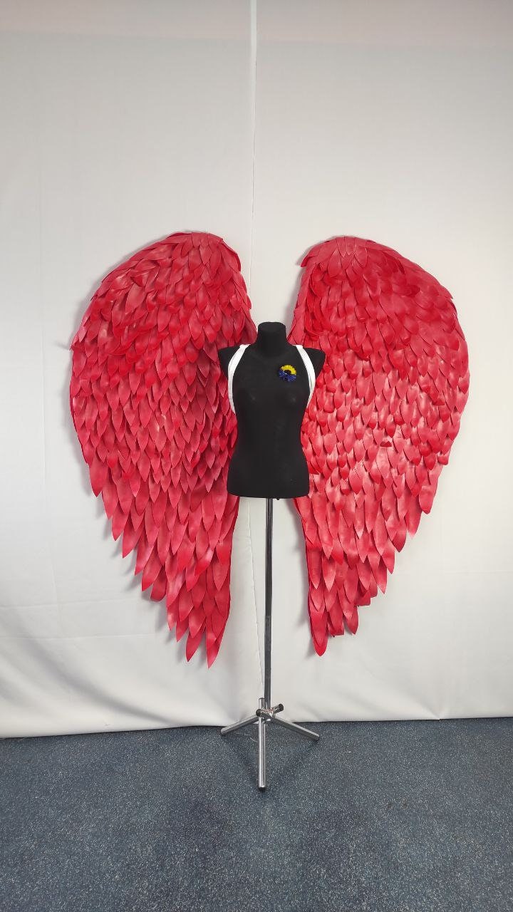 May include: Large, vibrant red angel wings with a detailed feather design. The wings are displayed behind a black mannequin with a white harness and a small, colorful emblem. The wings are on a metal stand against a white backdrop.
