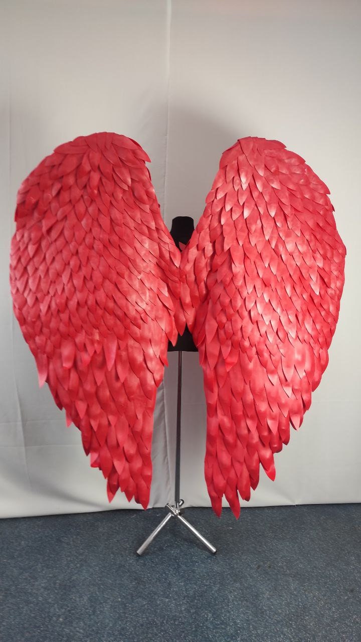 May include: Large, vibrant red angel wings, constructed from layered, feather-like material. The wings are displayed on a black mannequin torso, mounted on a silver stand, against a white backdrop. The wings are a striking costume piece.