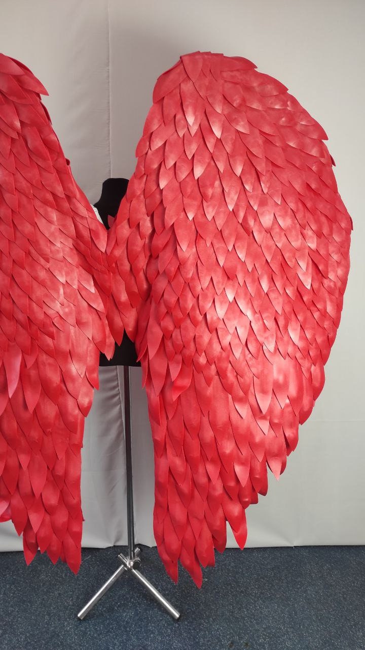 May include: A pair of large, vibrant red angel wings. The wings are constructed from numerous overlapping, leaf-shaped pieces, creating a textured, feathered appearance. They are displayed on a black mannequin form against a neutral backdrop.
