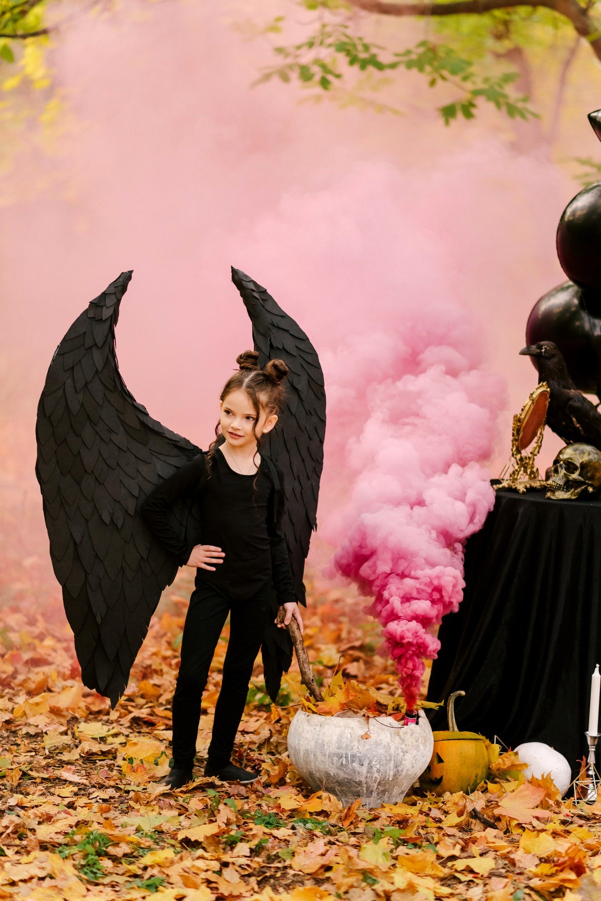 May include: A young girl dressed as a witch with black wings stands in a forest setting. She holds a wooden stick in her hand and is surrounded by pink smoke. There is a cauldron in front of her and fall leaves on the ground.
