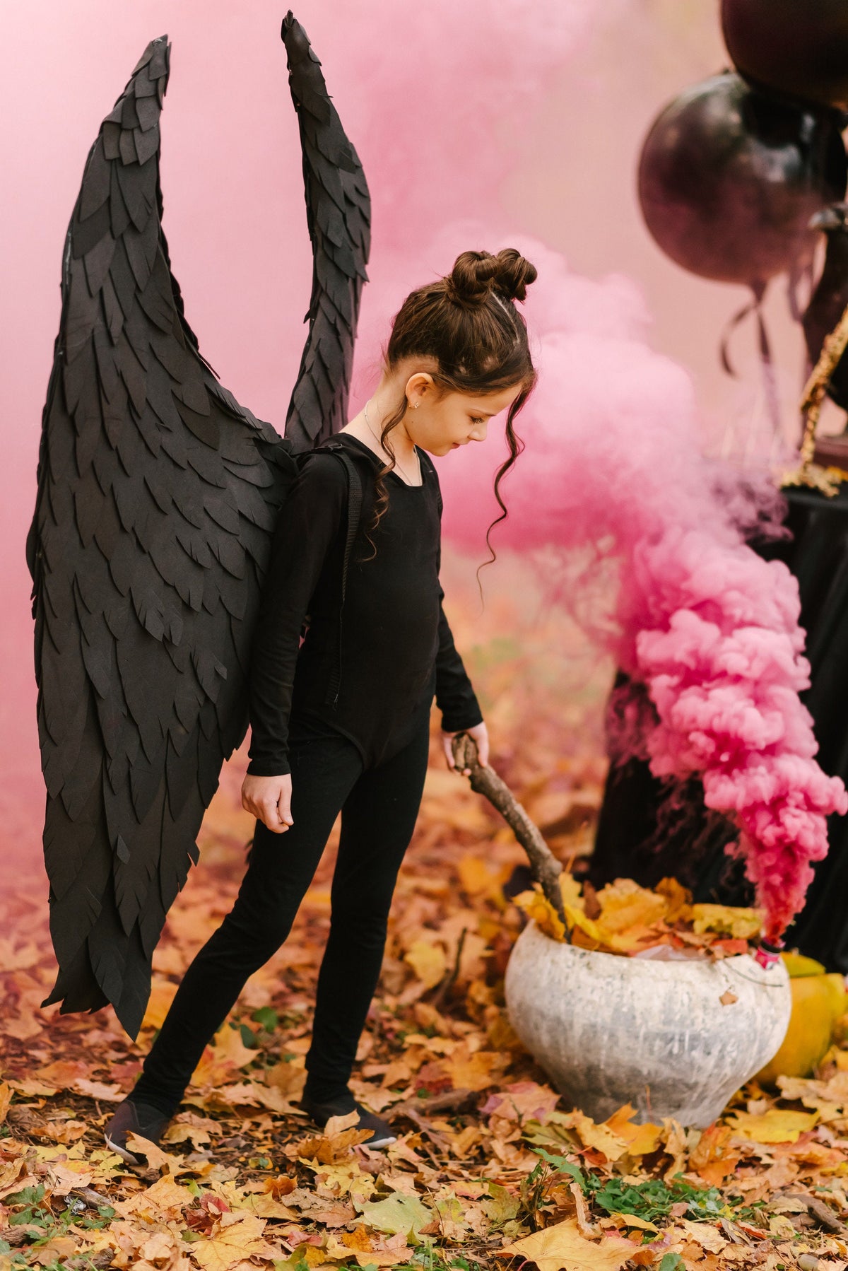 May include: A young person dressed in black with large black wings stands in a forest setting. They are holding a stick and looking down at a cauldron filled with leaves. Pink smoke billows behind them.