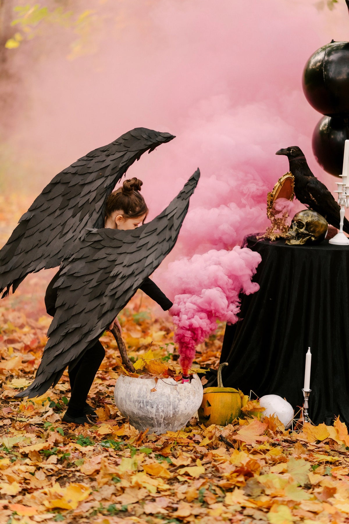 May include: A young girl dressed as a raven with black wings stands in a forest setting. She is holding a stick and stirring a cauldron. Pink smoke billows around her. There are pumpkins and other Halloween decorations in the scene.