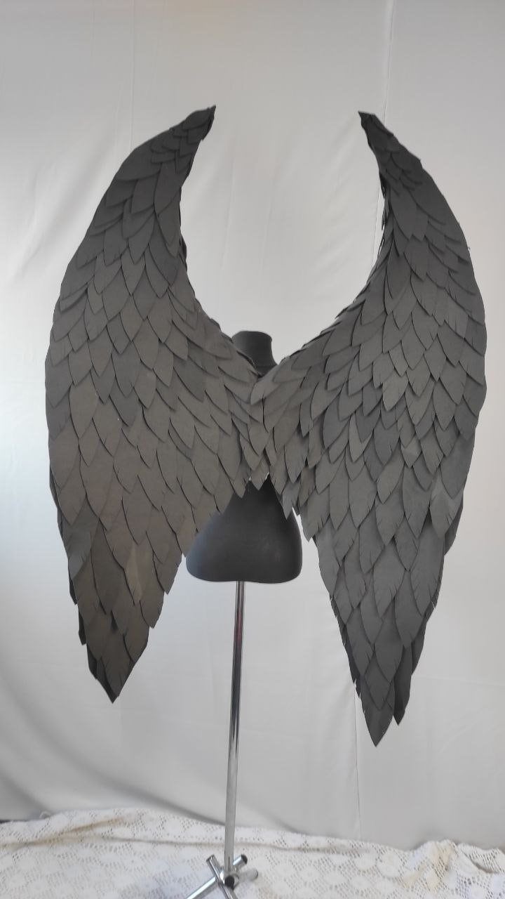 May include: A pair of large, black, feathered angel wings. The wings are made of individual feathers that are layered and attached to a wire frame. The wings are displayed on a mannequin stand.