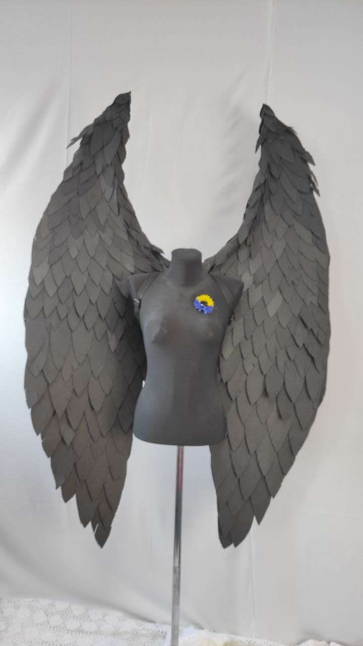 May include: A pair of large, black, feathered angel wings on a mannequin. The wings are made of individual feathers that are layered and overlapping. The wings are attached to the mannequin's back with straps.