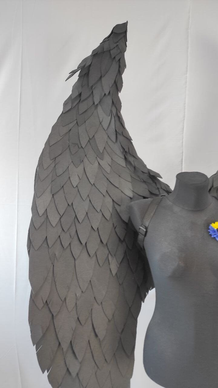 May include: A pair of black angel wings made of layered paper feathers. The wings are attached to a black mannequin torso.