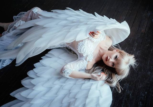 May include: A woman wearing a white dress with large, feathered angel wings. The wings are spread out, and the woman is lying on a dark wooden floor. The dress has lace detailing, and the woman has blonde hair and red lipstick.