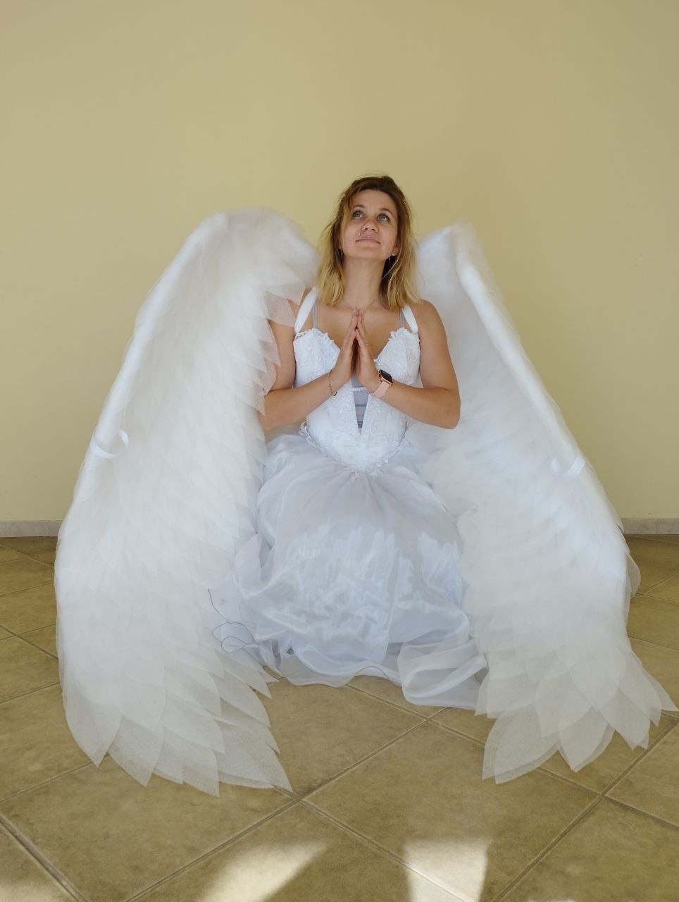 May include: A person wearing a white dress and large white angel wings, kneeling with hands clasped in prayer. The wings are large and feathered, and the dress is detailed with lace and tulle. The background is a plain, light-colored wall.