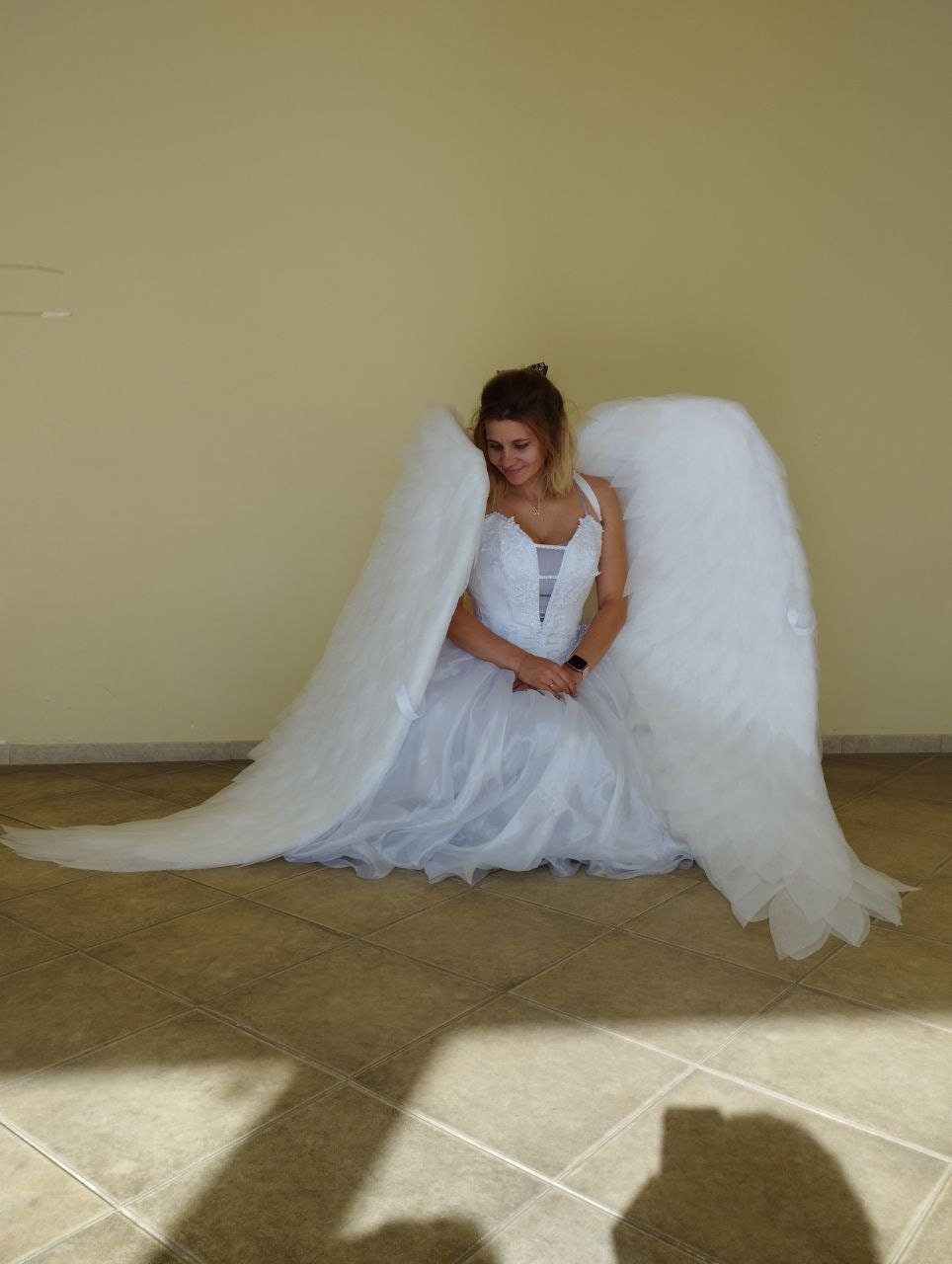 May include: A person wearing a white wedding dress and large white angel wings, kneeling on a tiled floor. The dress has a fitted bodice and a flowing skirt. The wings are large and feathery.