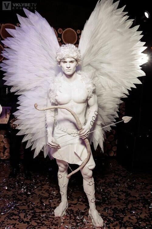 May include: A person dressed as Cupid with white feathered wings, a white loincloth, and a bow and arrow. The person is standing on a floor covered in confetti.
