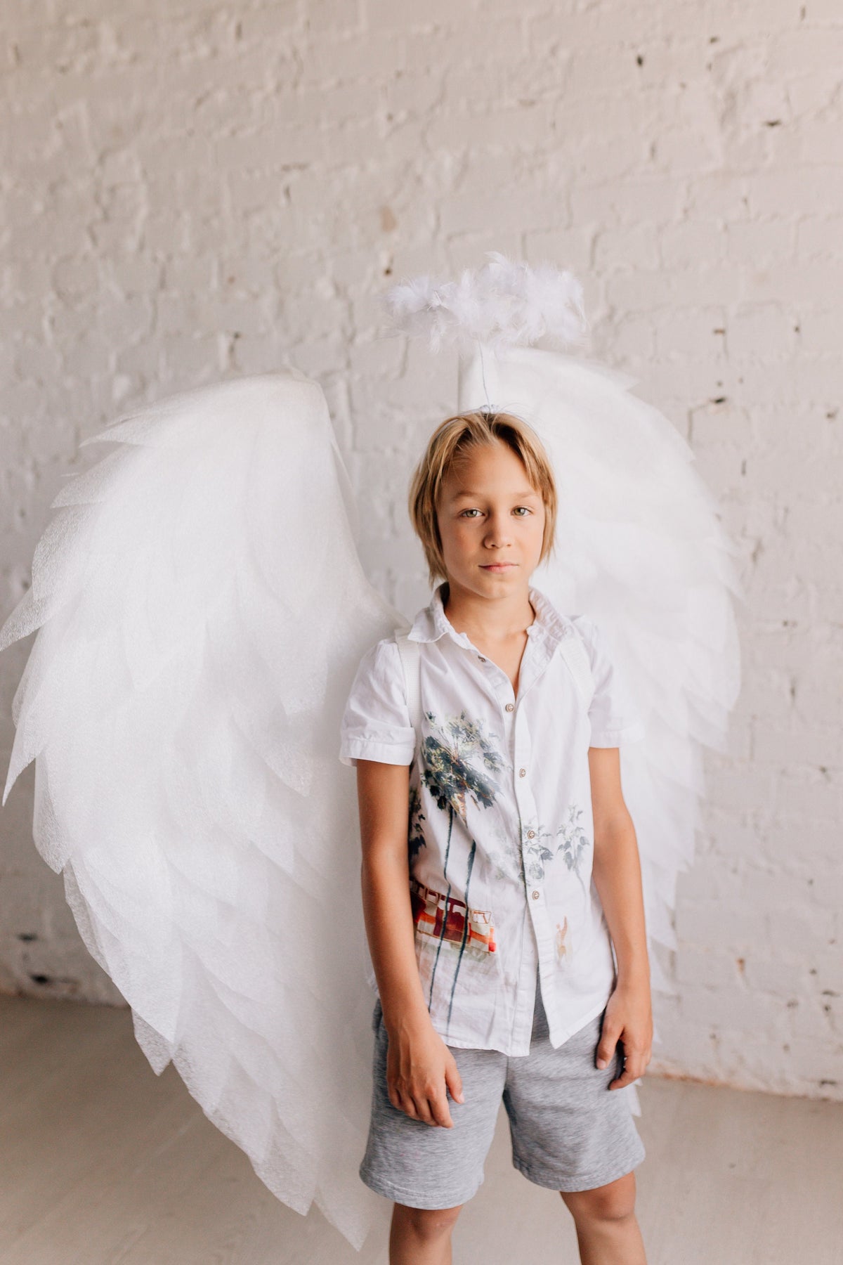 May include: A young person wearing a white shirt with a palm tree print and gray shorts. They are wearing a white angel wing costume with a halo headband.