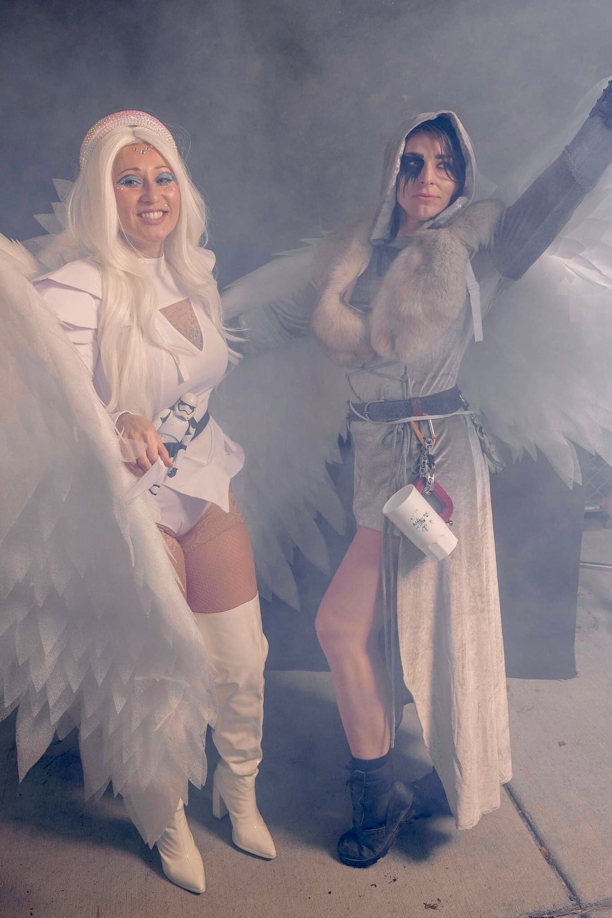 May include: Two people dressed as angels, one in white with a white feathered halo and white boots, the other in grey with a fur collar and black boots. Both are holding white feathered wings.