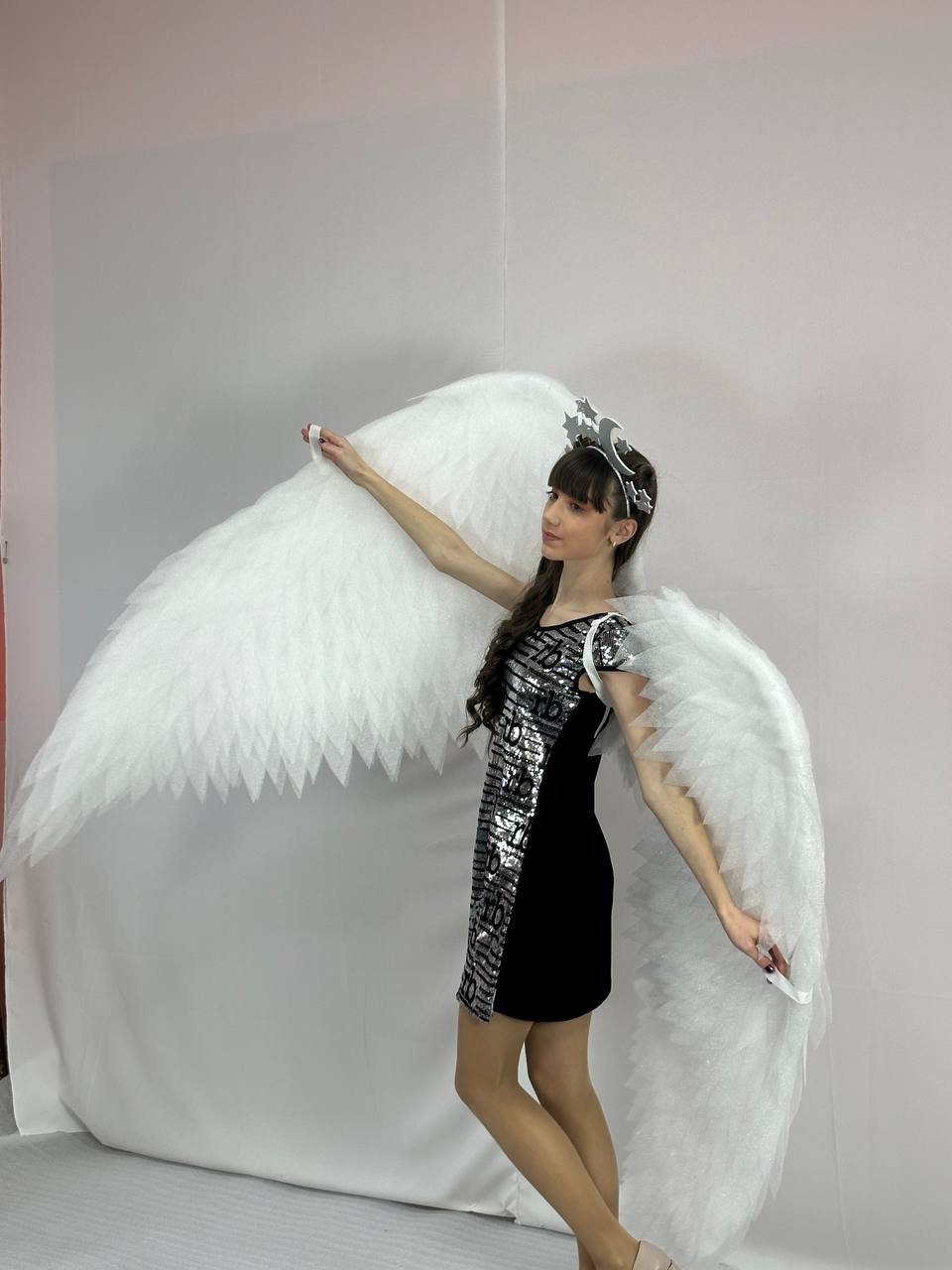May include: A woman wearing a black dress with silver sequins and a silver headband with stars and a crescent moon. She is holding large white feathered angel wings.