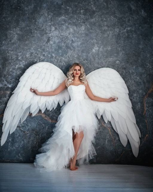 May include: A woman wearing a white dress with a long train and large white angel wings. She is standing in front of a gray textured wall.