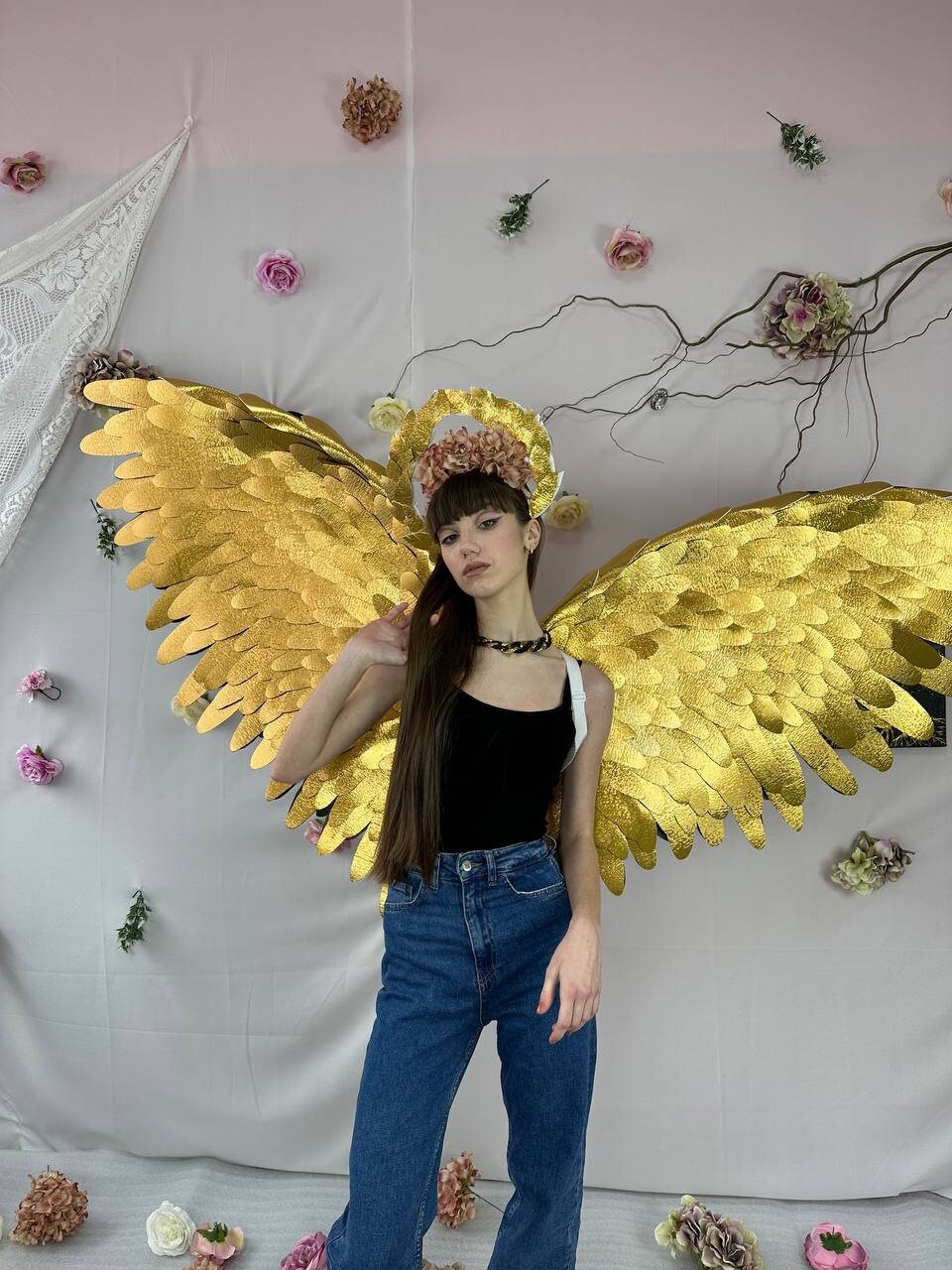 May include: A person wearing a black tank top and blue jeans with gold angel wings and a flower crown.