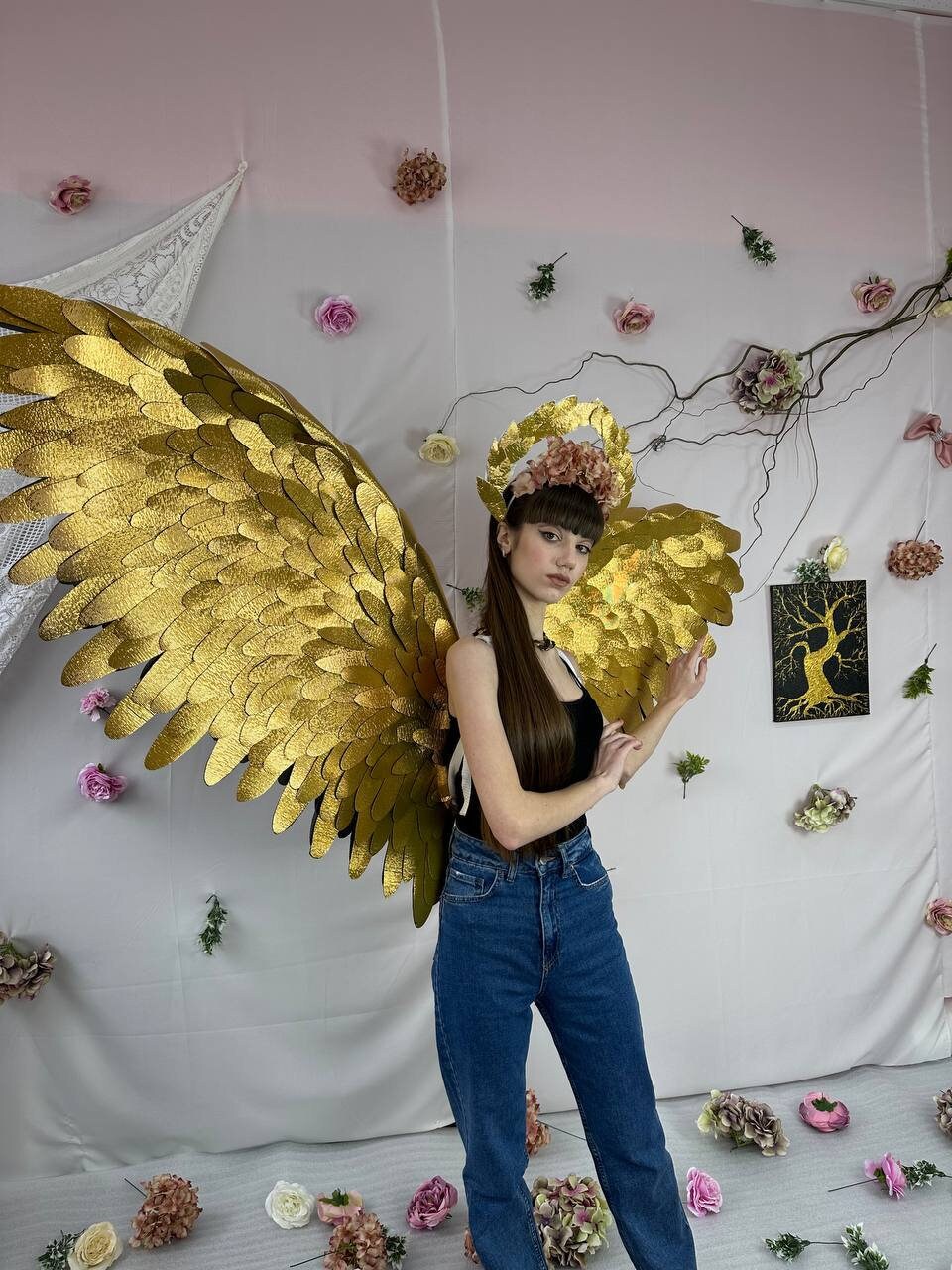 May include: A person wearing a black top and blue jeans with large gold wings and a floral crown. The background is a pink and white wall with a vine of flowers.