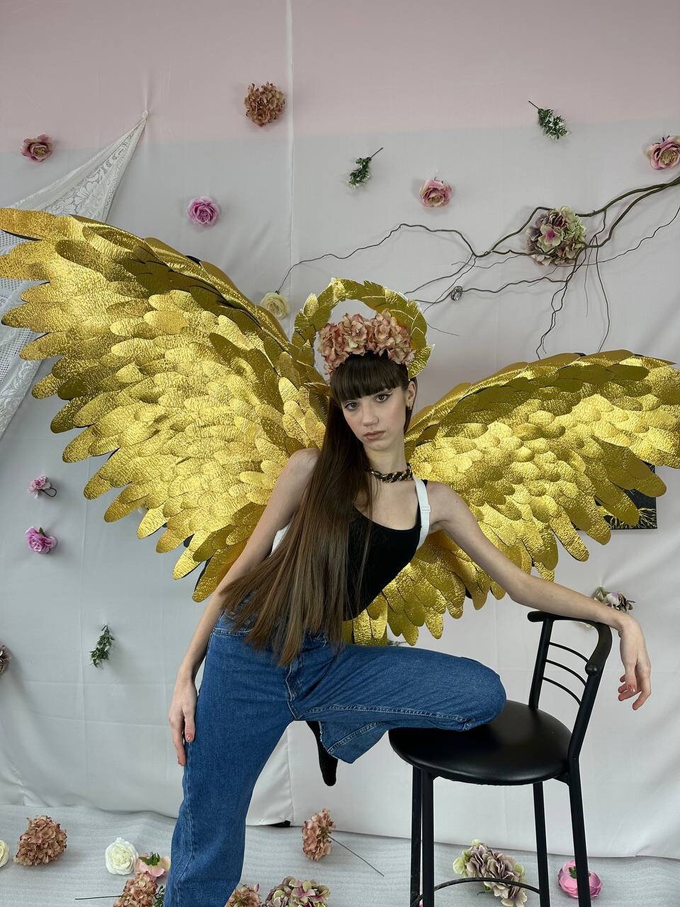 May include: A person wearing a black tank top and blue jeans poses with large gold angel wings and a flower crown. The background is a pink and white wall with a white lace curtain and a branch with pink flowers.
