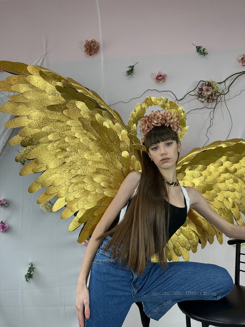 May include: A person wearing a black tank top and blue jeans with large gold angel wings and a flower crown.