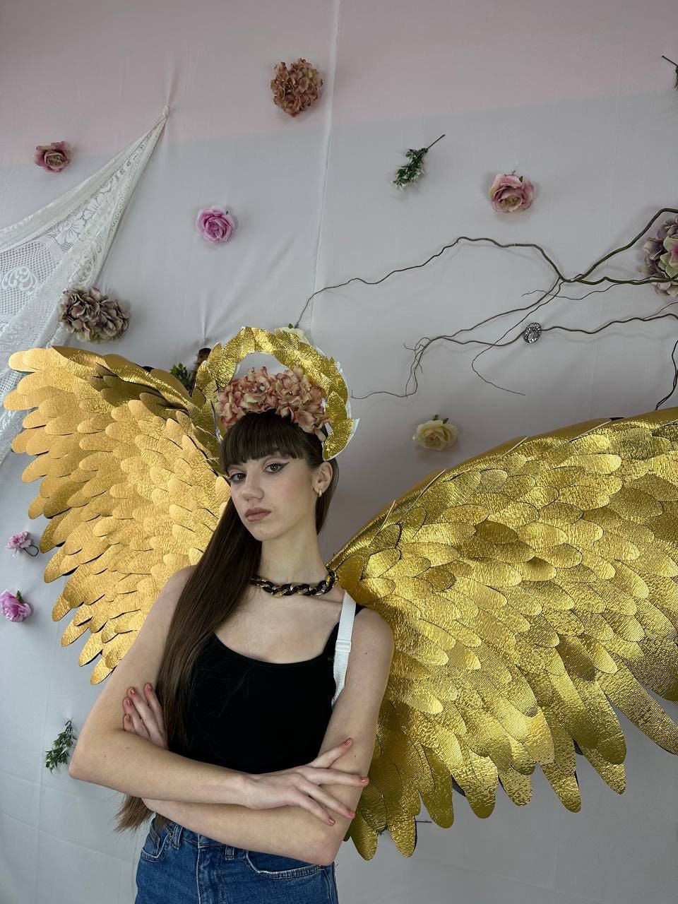 May include: A person wearing a black tank top and blue jeans with gold angel wings and a floral crown.