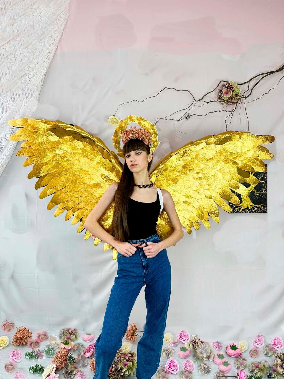 May include: A person wearing a black tank top and blue jeans with large gold angel wings and a flower crown.