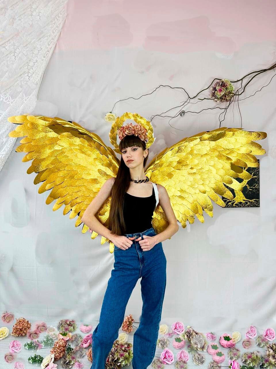May include: A person wearing a black tank top and blue jeans with large gold angel wings and a flower crown.