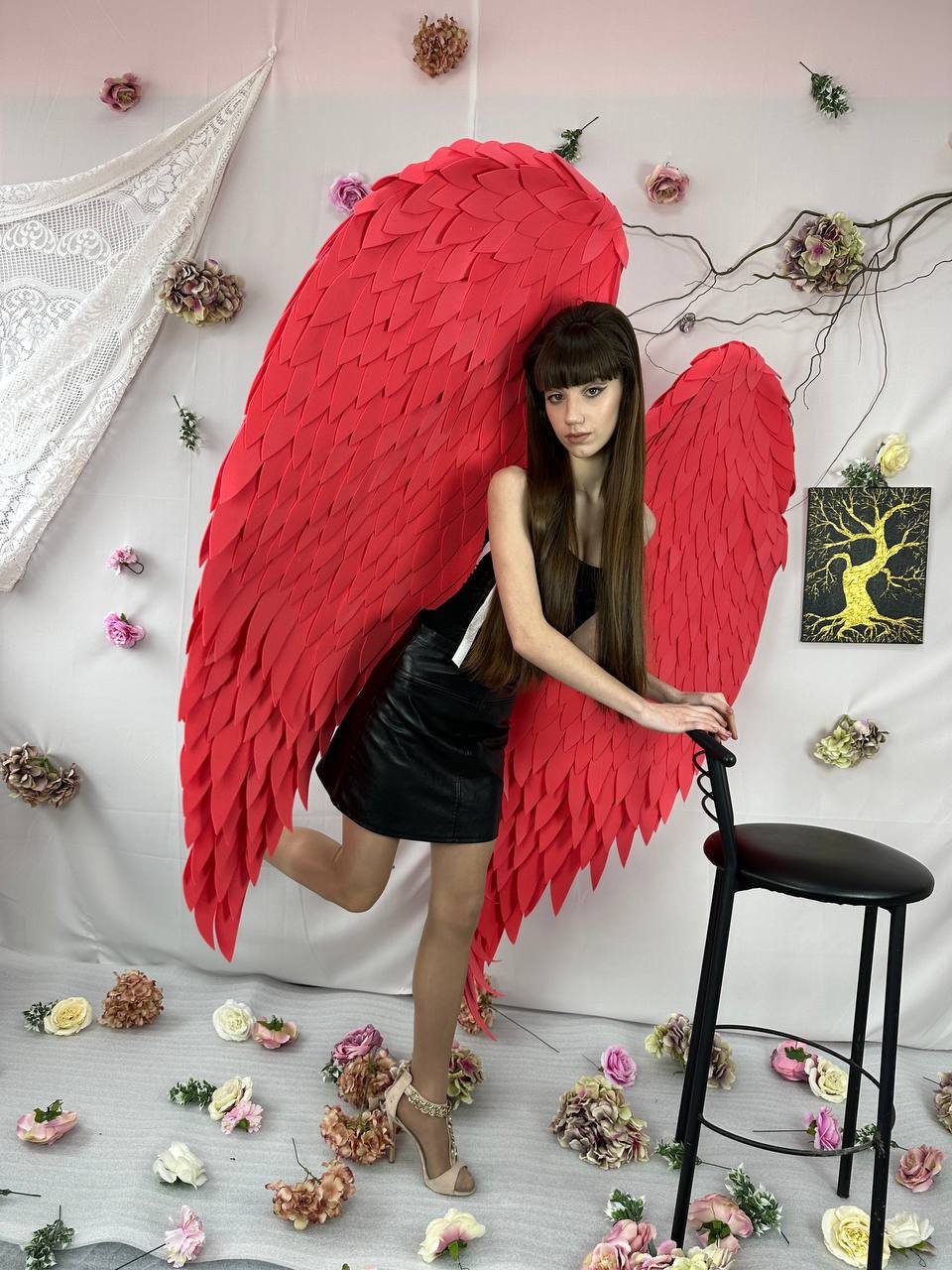 May include: A person wearing large, vibrant red angel wings. The wings are made of layered, textured material. The person is wearing a black top and skirt, with high-heeled shoes. A black stool is nearby, and flowers are scattered around.