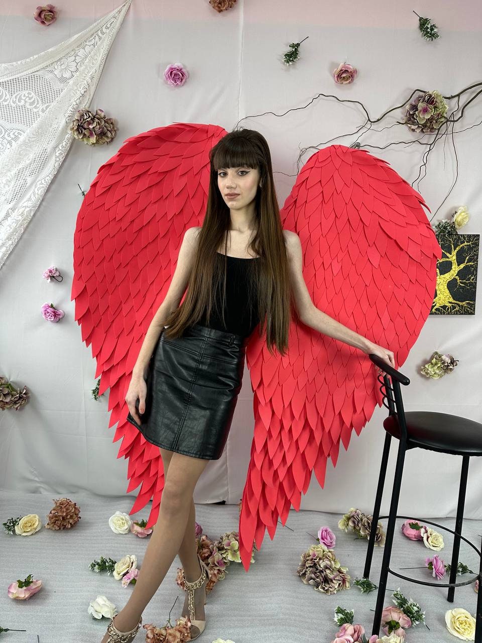 May include: A woman poses with large, vibrant red angel wings. She wears a black top, a black leather skirt, and beige heels. The background includes floral decorations and a black chair.