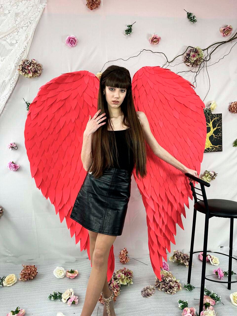 May include: A person wearing a black top and skirt with large, bright red angel wings. The wings are made of feathers and are attached to the person's back.