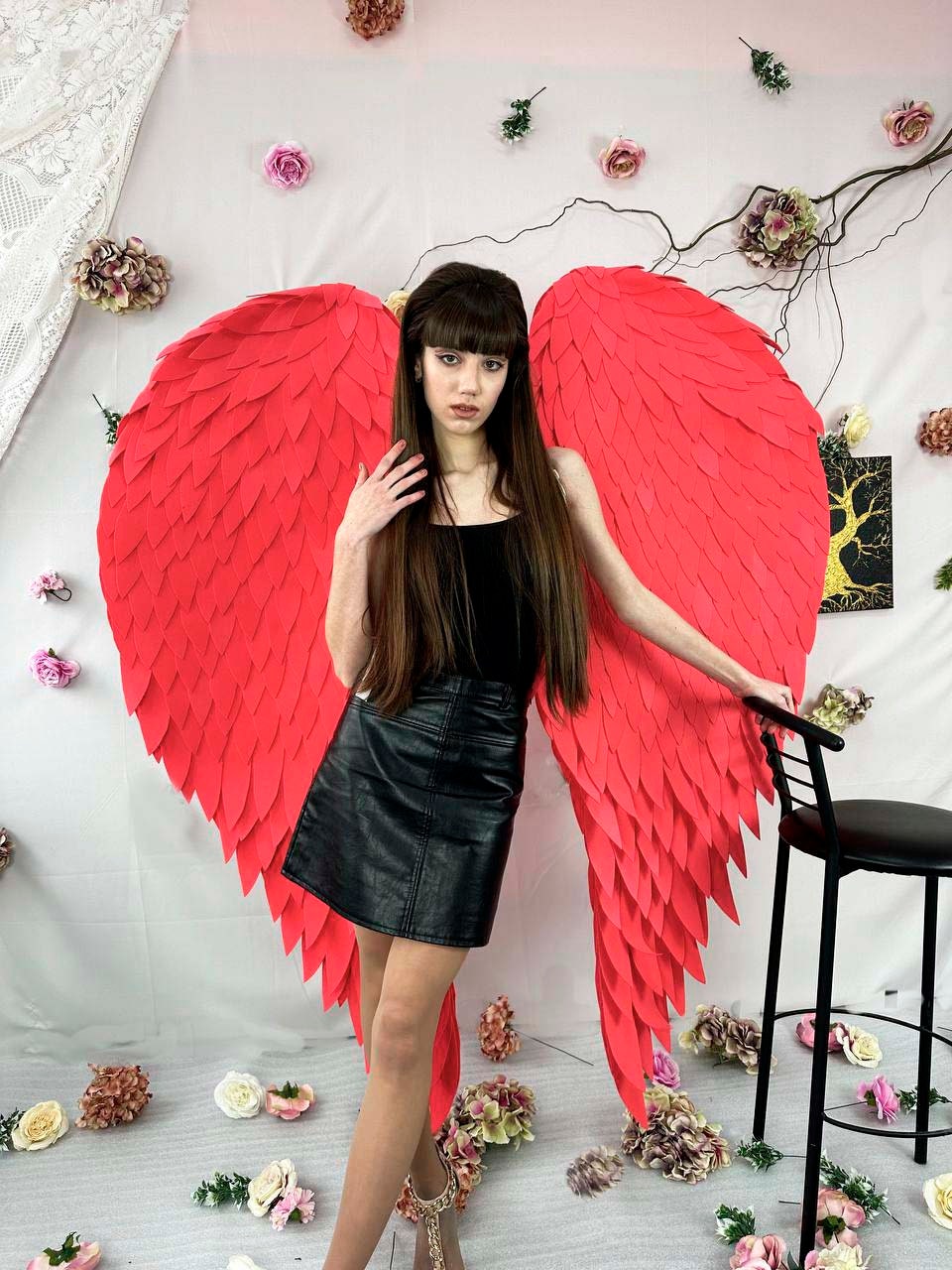 May include: A person wearing a black top and a black leather skirt stands in front of large, vibrant red angel wings. The wings are the focal point, with a textured, layered design. The background includes floral decorations and a black chair.