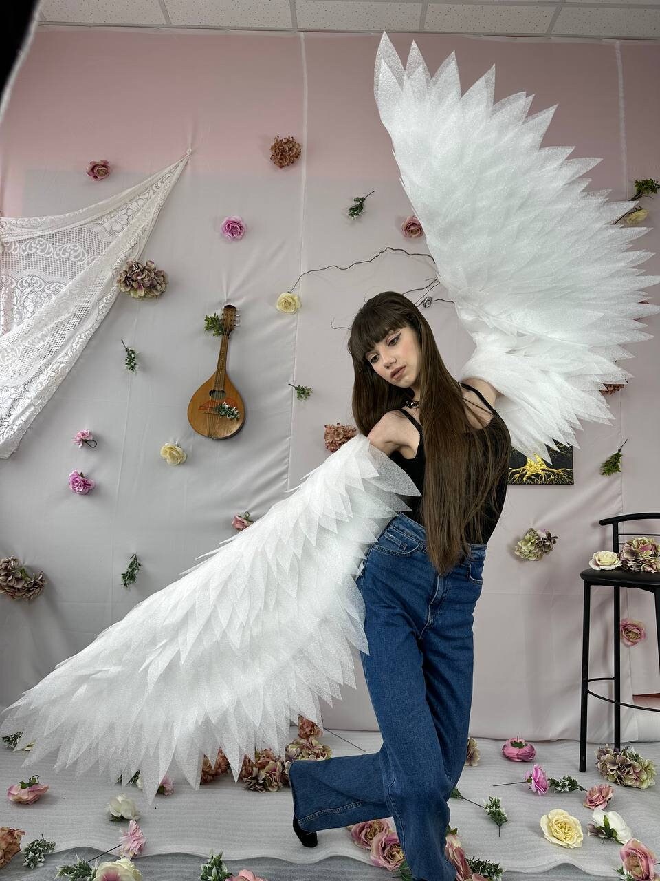 May include: A woman wearing a black tank top and blue jeans poses with large white feathered angel wings. The wings are made of many layers of white feathers and are very detailed. The woman is standing in front of a pink wall with white flowers and a white lace curtain.