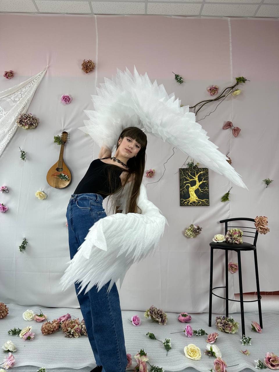 May include: A person wearing a black top and blue jeans is holding large white feathered wings. The wings are spread out and the person is looking at the camera. The background is a pink wall with white flowers and a small wooden chair.