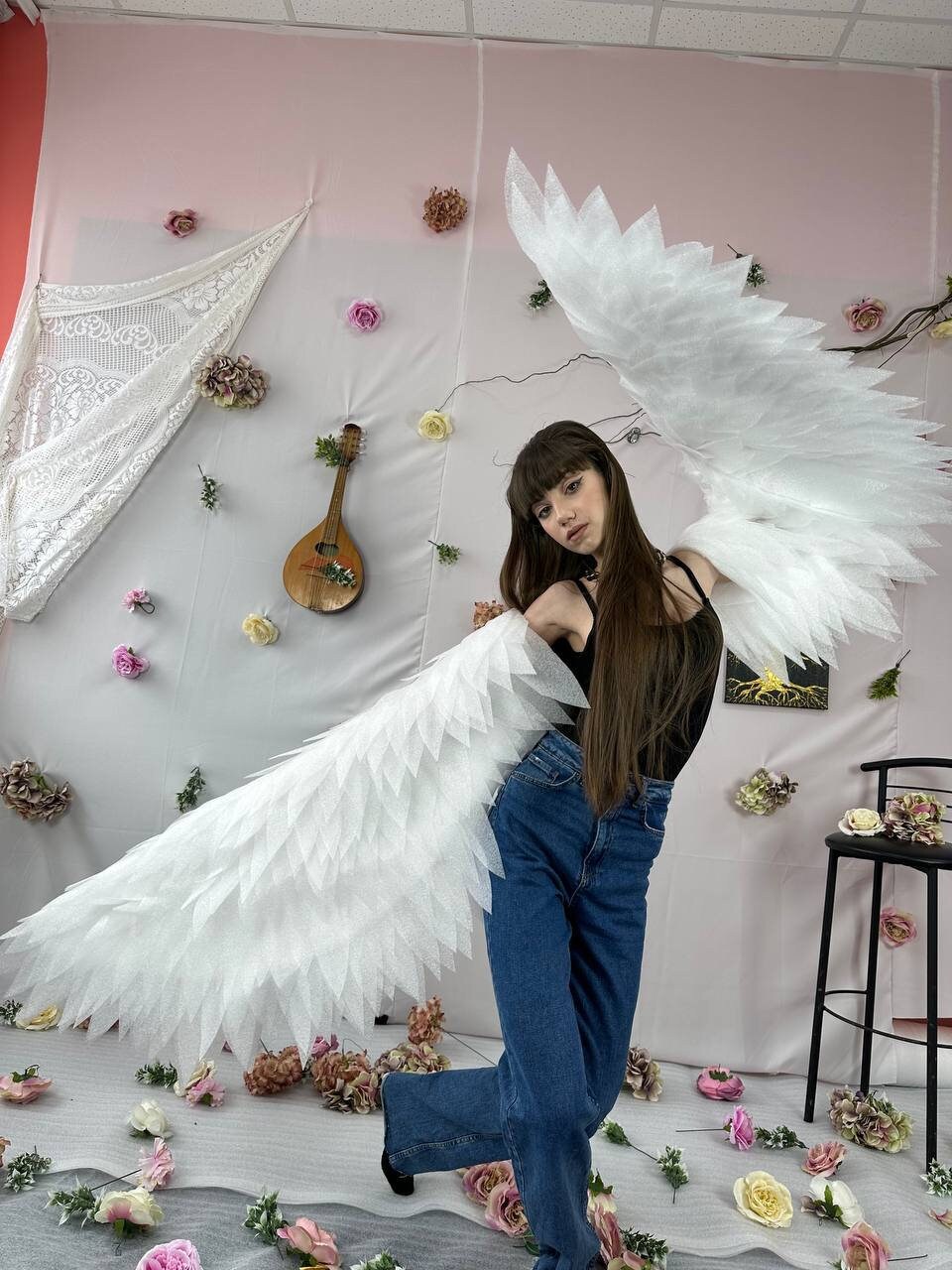 May include: A person wearing a black tank top and blue jeans poses with large white angel wings. The wings are made of feathers and are very detailed. The person is standing in front of a pink and white wall with flowers and a guitar.