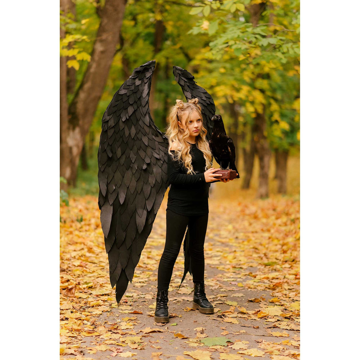 May include: A young person wearing black clothing and large black feathered wings, holding a black raven in their hands. The person is standing on a path in a forest with fallen autumn leaves.