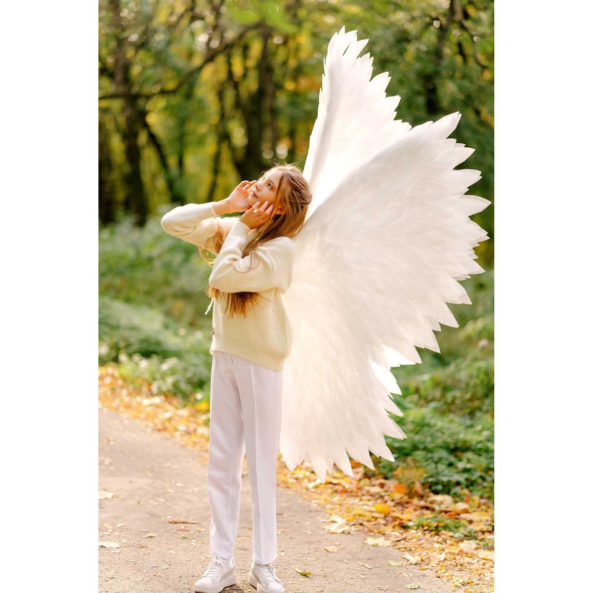 May include: A young person wearing a white sweater and white pants with large white angel wings. The person is standing in a forest setting with fall foliage on the ground.