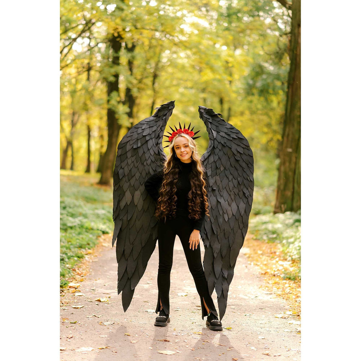 May include: A young person wearing a black outfit with black angel wings and a red flower crown. The person is standing on a path in a forest with fall foliage.