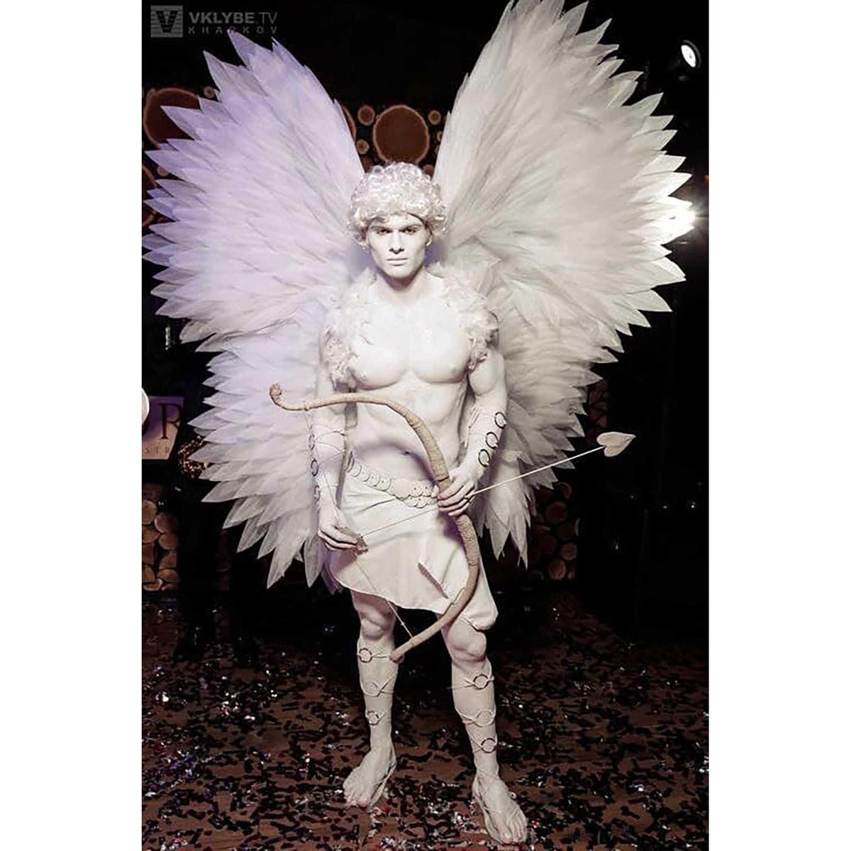 May include: A white-painted person dressed as Cupid, wearing a white wig, white wings, and a white loincloth. They are holding a bow and arrow.