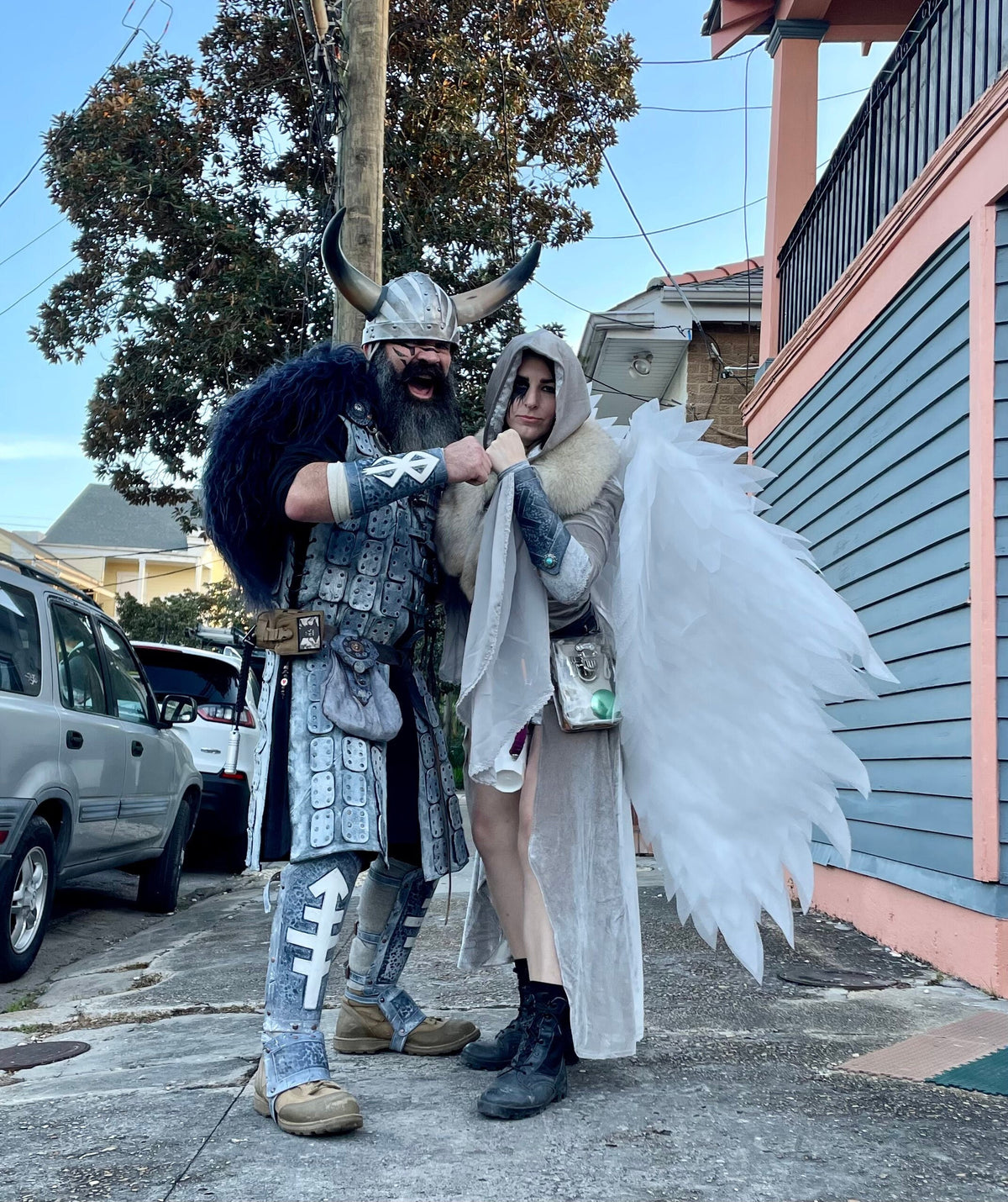 May include: A person dressed as a Viking warrior with a horned helmet and a fur-trimmed coat stands next to a person dressed as an angel with white wings. The angel is wearing a white dress and a fur-trimmed coat.